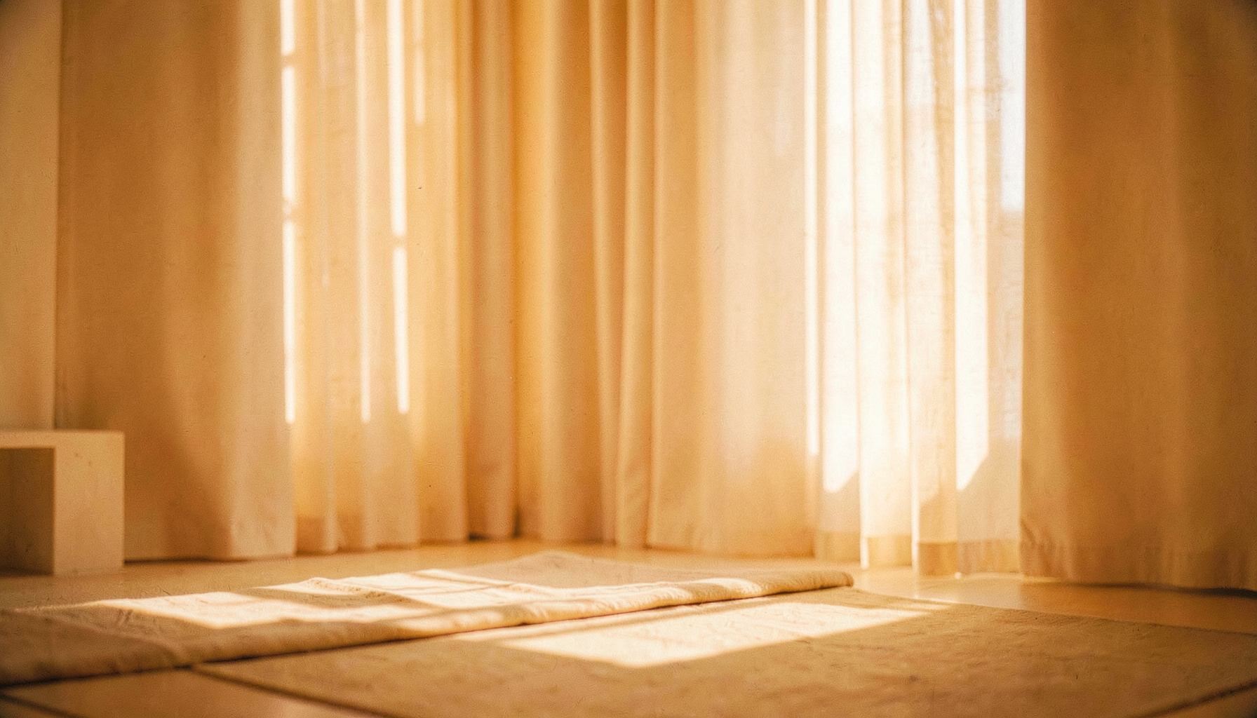 Soft warm light filtering through sheer curtains into a quiet meditation space, cream and sand tones with gentle shadows